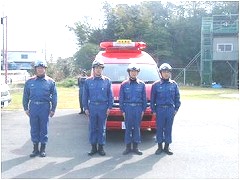 赤い消防車両の前に、ヘルメットを被り消防活動服を着用した4人の方が横一列に整列している写真