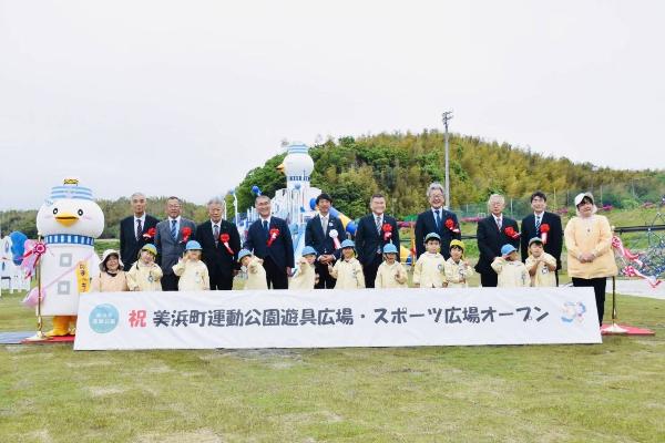 美浜町運動公園遊具広場・スポーツ広場のオープン記念式典において、伊藤衆議院議員、横田県議会議員をはじめとする来賓と町長、奥田保育所の園児、のまっキーが遊具広場を背に写真に写る様子