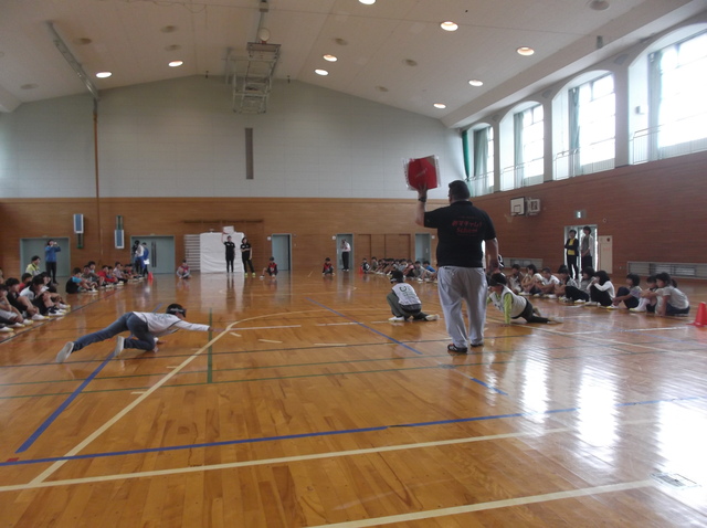 体育館の両端に児童が並んで座り、その間で体を床にはわせながらゴールボールの体験をしている児童3名と男性の写真