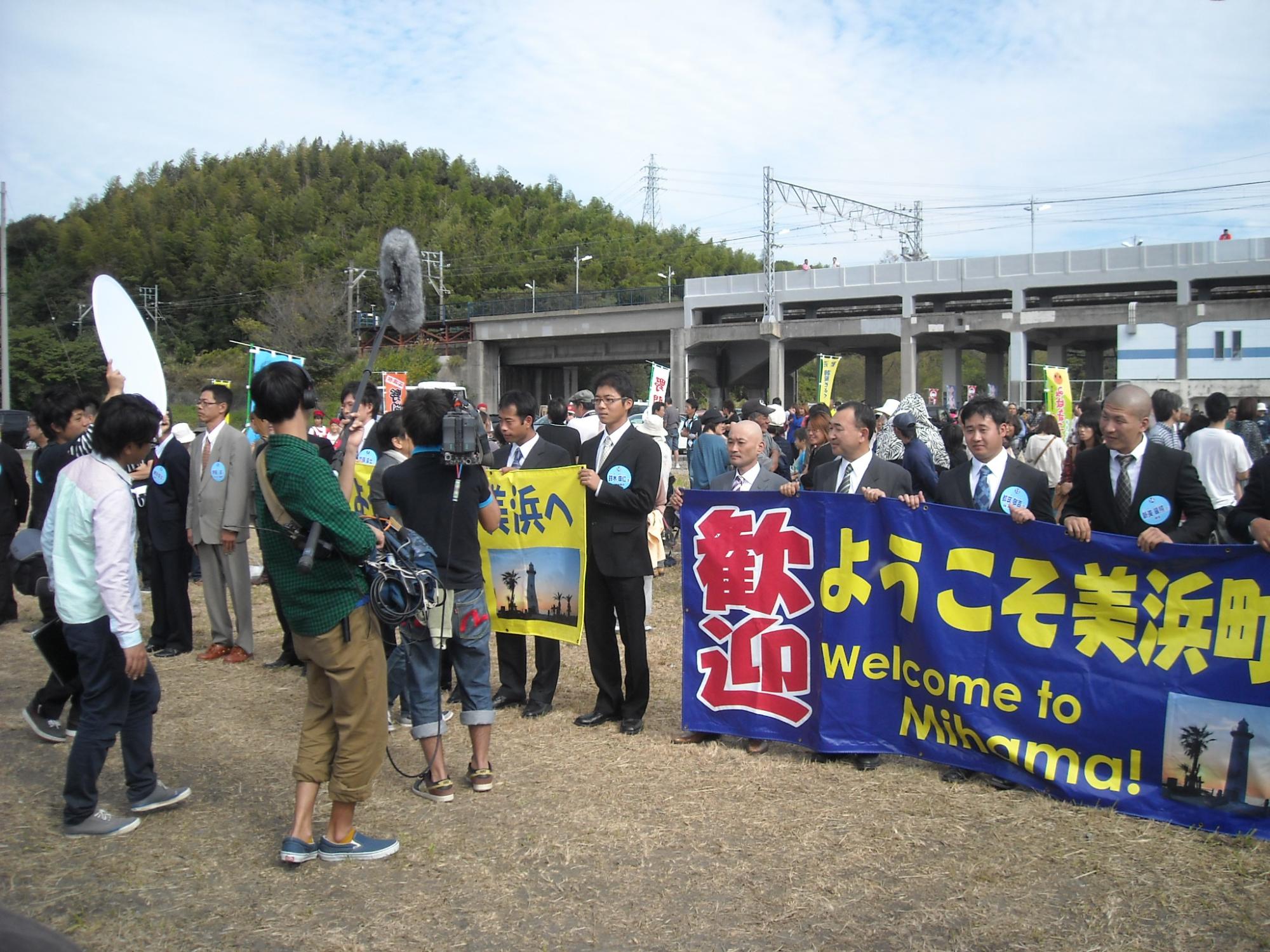 海辺に歓迎 ようこそ美浜町と書かれた横断幕の後ろに男性が並んでいる所をテレビカメラが撮影している様子の写真