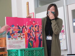 国際交流員の女性が台に置かれた大きな絵本を読みながら笑っている写真