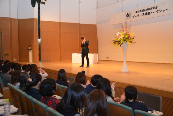 舞台の中央に花が置かれゲッターズ飯田さんが話しているのを沢山の人が客席で聞いている写真