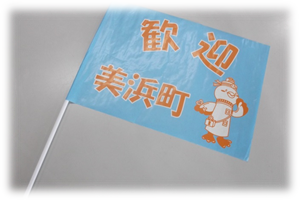 水色の旗にオレンジ色で歓迎 美浜町と書かれ、のまっキーのご当地キャラクターが描かれた手振り旗