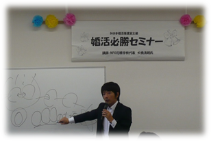 「婚活必勝セミナー」と書かれた横断幕の下、マイクを持った男性がホワイトボードに書きながら話している写真
