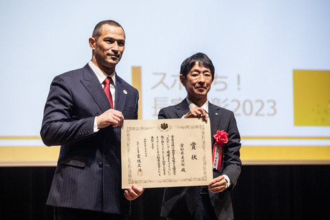 大きなスクリーンの前で、室伏広治スポーツ庁長官と町長が賞状を一緒に持ち、記念撮影をしている写真