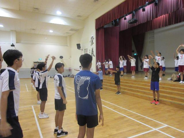 ニーアン中学校の体育館で、派遣された生徒が壇上でラジオ体操を披露し、ニーアン中学校の生徒がそれを見ながら体操する様子。