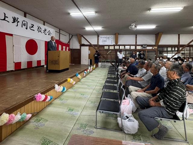 上野間区の敬老会で、区長が壇上で参加者に話している様子。
