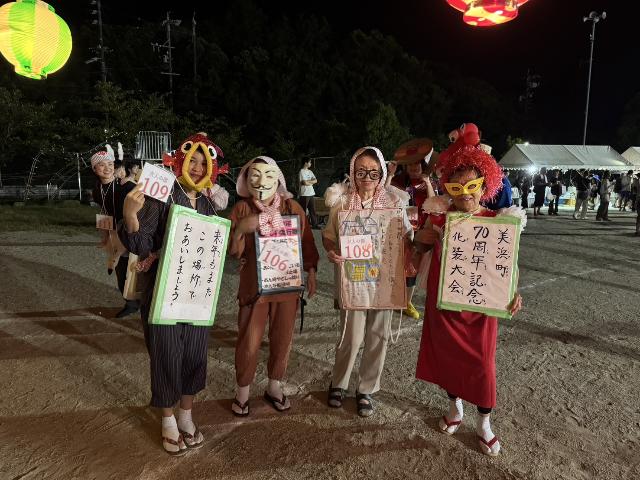 2025年の奥田地区の盆踊りで仮装した4人がパネルを持ち並んで写っている様子