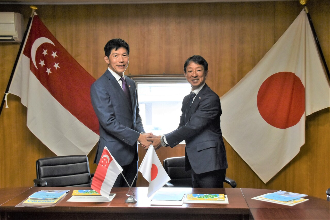 シンガポールと日本の国旗が飾られた部屋で、駐日大使と町長が握手をし記念撮影をしている写真