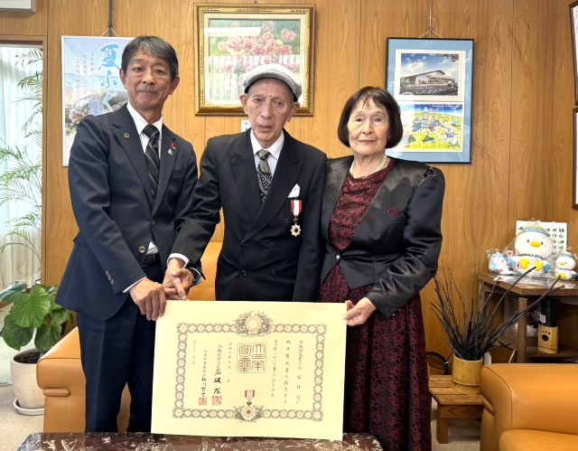 旭日雙光章を叙勲された平塚さんが町長へ表敬訪問し町長と奥様と賞状をもって並んで写真を撮っている様子