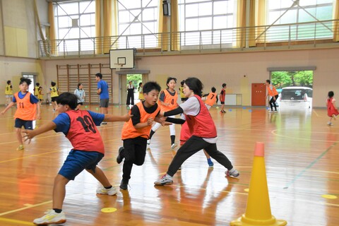 河和小学校体育館で、赤いゼッケンを付けた男の子と女性がオレンジのゼッケンを付けた男の子を捕まえようとしているスポーツ鬼ごっこの写真