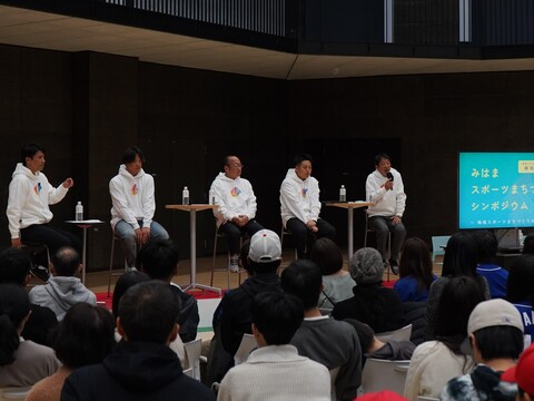 体育館で、男性が椅子に座りマイクを持って話をし、参加者の方々が白い椅子に座って聞いているシンポジウムの様子の写真