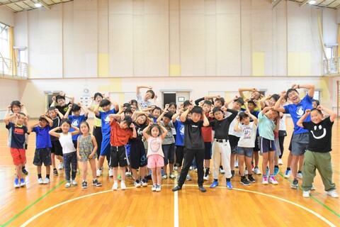 河和小学校体育館で、参加者の子供たちと大人が両手で拳を作り頭に当てたポーズをとっている集合写真