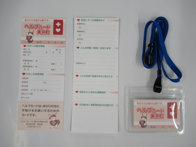 「ヘルプカード美浜町」と書かれている長方形をした氏名や生年月日等を記載する項目のあるヘルプカード表と裏、青い紐の付いたネームタグに入っているヘルプカードのが並べて置かれている写真