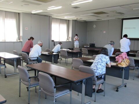 スクリーンがある室内で、2列に並べられた長机に椅子が2脚ずつ設置された席で参観者たちが中腰になり、両手を胸の部分でクロスして体操を行っている様子の写真