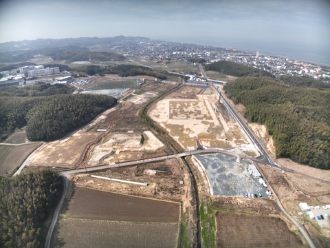 周囲に民家や鬱蒼と茂った森が点在する陸上競技場の建設予定地で、観覧スタンドの基盤設備工事が進められている様子を令和4（2022）年3月斜め上空から撮影した写真