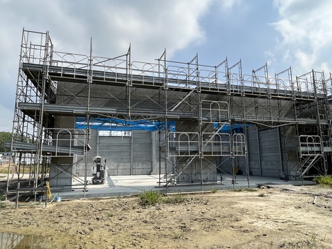 周囲に足場が設置され、建物内の天井に青色のネットが設置された陸上競技場器具庫の壁の設置工事の様子を正面側から撮影した写真