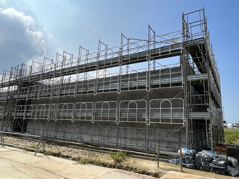建物の外側に資材が並んで置かれ、足場が設置された陸上競技場器具庫の壁を設置している工事の様子を建物の傍から撮影した写真
