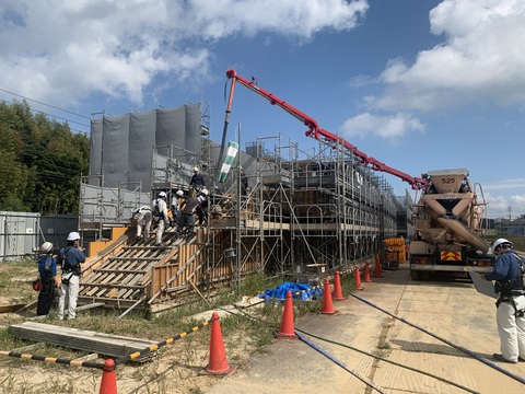 三角コーンで囲まれた建設中の陸上競技場の階段部分まで長いブームが伸びて、7名の作業員がコンクリートを打設工事を行っている様子の写真