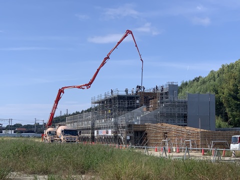 建設中の陸上競技場3階部分に、建設現場の前に停車しているコンクリートポンプ車から延びた長いブームでコンクリートを打設が行われている様子の写真