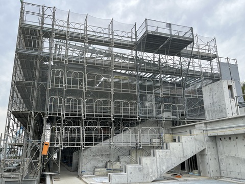 周囲に足場が組まれネットで覆われた陸上競技場観覧スタンドの屋根部分工事が行われている様子を側面から撮影した写真