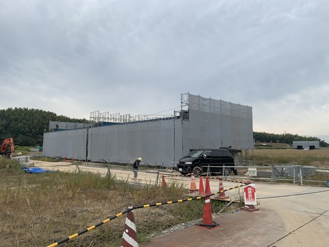 周囲に足場が組まれネットで覆われた陸上競技場器具庫の屋根取付工事が行われている様子の写真