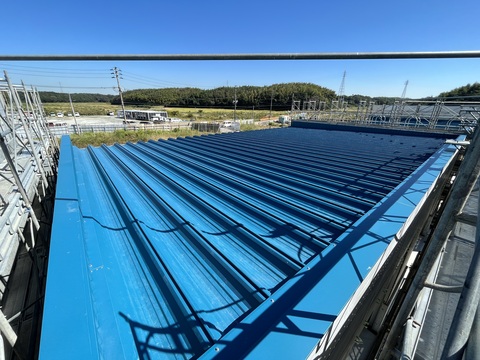 周囲に足場が組まれた陸上競技場器具庫の屋根に水色の屋根の取付工事が行われた様子を撮影した写真
