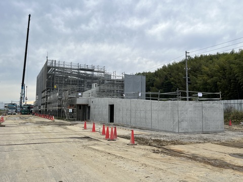 建設現場の前に三角コーンが並び、足場が組まれている陸上競技場観覧スタンドの傍に停車しているクレーン車から大きなアームが伸びて工事が行われている様子の写真