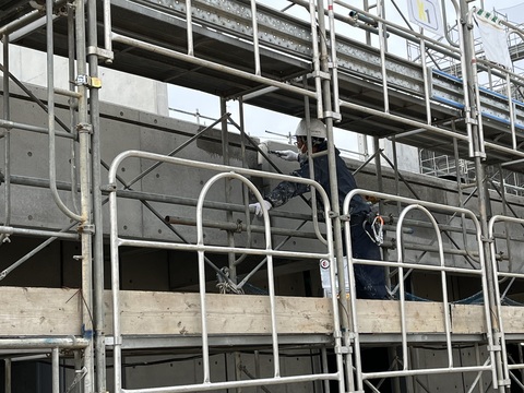周囲に足場が組まれた陸上競技場観覧スタンド外壁部分を、ヘルメットを被った作業員が塗装工事を行っている様子の写真