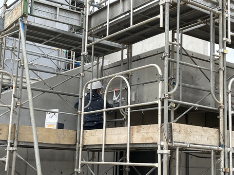 陸上競技場観覧スタンドの周囲に組まれた足場にヘルメットを被り座っている作業員が、塗装工事を行っている様子の写真