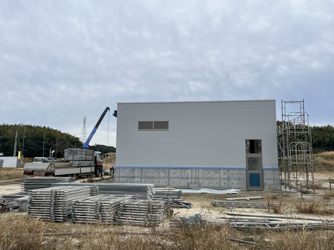 白壁に塗装され、右下に出入り口が設置された陸上競技場器具庫の側面の足場が撤去され、手前に解体した足場が種類ごとに並べて置かれている写真
