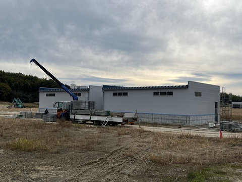 白壁に塗装された陸上競技場器具庫の傍に停車したトラックの荷台に、解体した足場がクレーンで積まれている様子の写真