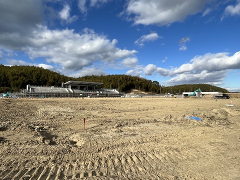 鬱蒼と茂った森の手前に建設中の陸上競技場があり、手前の地面に位置を示す杭がささっているフィールド部分の基盤整備工事の様子写真