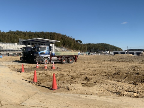 建設中の器具庫や工事車両がある左手前に三角コーンが置かれ、2台のダンプカーが停車している、陸上競技場フィールド部分の基盤整備工事が行われている様子の写真