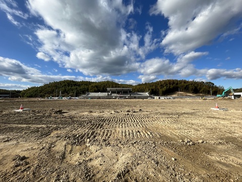鬱蒼と茂った森の手前に建設中の陸上競技場があり、手前左右の地面に位置を示す三角コーンが置かれている、フィールド部分の基盤整備工事の様子写真