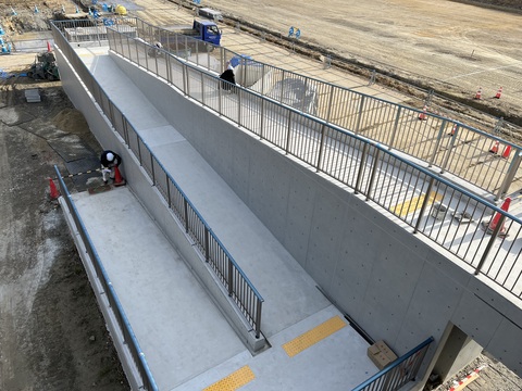 3名の作業員が陸上競技場観覧スタンドスロープ部分で手すり設置工事を行っている様子の写真