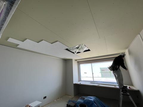 ヘルメットを被った作業員が脚立に乗りながら、天井にボードを貼り付けている工事の様子写真