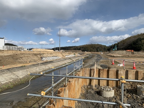川沿いの奥の盛り土の上で工事車両が工事を行っている様子を、人道橋の橋台部分の基礎工事側から撮影した写真