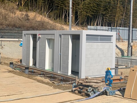 右側に資材が並べられ、左から男性トイレ、バリアフリートイレ、女子トイレが四角形の建物内に設置された屋外トイレの写真