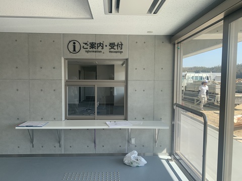 出入り口近くのコンクリート製の壁に設置された四角窓の上部に「ご案内・受付」と書かれたサインが取り付けられた写真