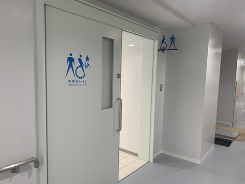 右上に縦長ですりガラスが施された引き戸の中央上部と室内札に、青色で人型のマークと車いすのマークのピクトグラム、オストメイト用とおむつ交換台のピクトグラム「男性用トイレRestroom for men」と書かれたサインが取り付けられた写真