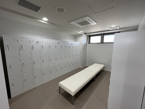 奥の壁上部に小窓が設置され、左右の壁に縦に3列の36個ずつロッカーが備え付けられ、中央に白色の2脚のベンチが設置されたロッカールームの写真