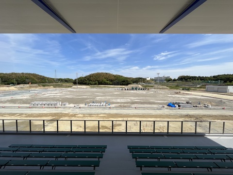 大きな屋根が設置された緑色の椅子の観覧席の後方から、建設中の陸上競技場のフィールド全体を撮影した写真