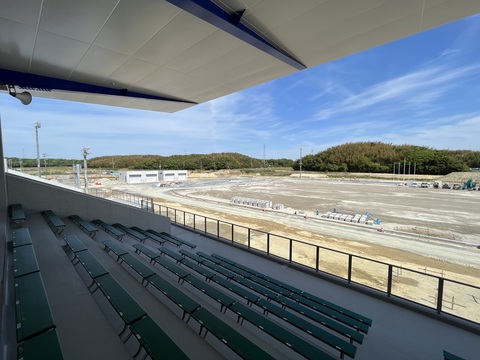 大きな屋根が設置された緑色の椅子の観覧席の後方から、建設中の陸上競技場のフィールドと器具庫のある左側を撮影した写真