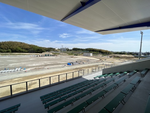 大きな屋根が設置された緑色の椅子の観覧席の後方から、建設中の陸上競技場のフィールドの右側を撮影した写真