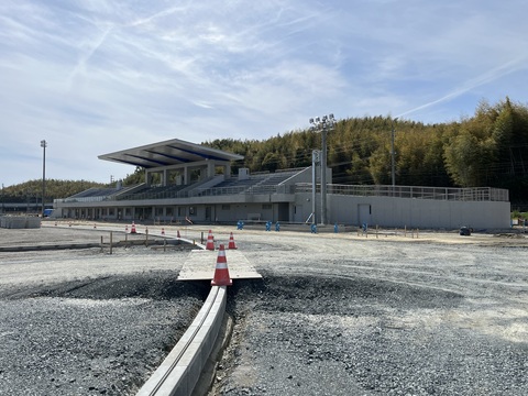 木が鬱蒼と茂った森の手前に建設された観覧スタンド全体を、建設中のフィールドから撮影した写真