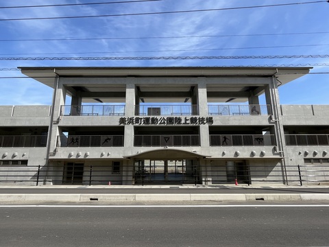 2階と3階の間の壁に「美浜町運動公園陸上競技場」と書かれた出入り口を、道路の反対側から撮影した写真