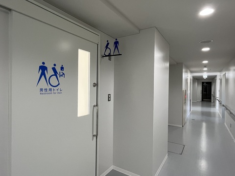 丸い照明が等間隔に天井に施された通路の左側に男性用トイレが設置されている写真