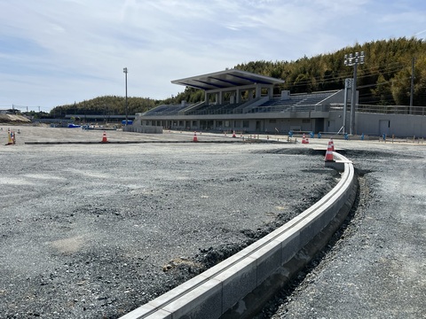完成した観覧スタンドの手前のフィールド沿いに三角コーンが置かれ、トラックとフィールドの間の部分にブロックが並び側溝が設置されている工事の様子写真