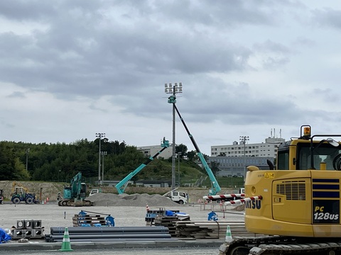 高所まで伸びた水色のボディのクレーン車2台で照明塔の電気配線工事を行っている様子の写真
